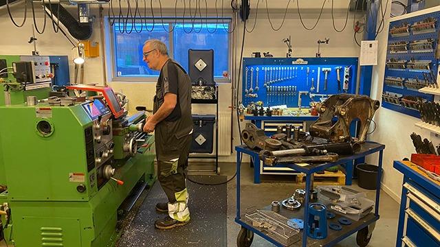 En mand arbejder ved en grøn drejebænk i et værksted med værktøjer og maskiner.