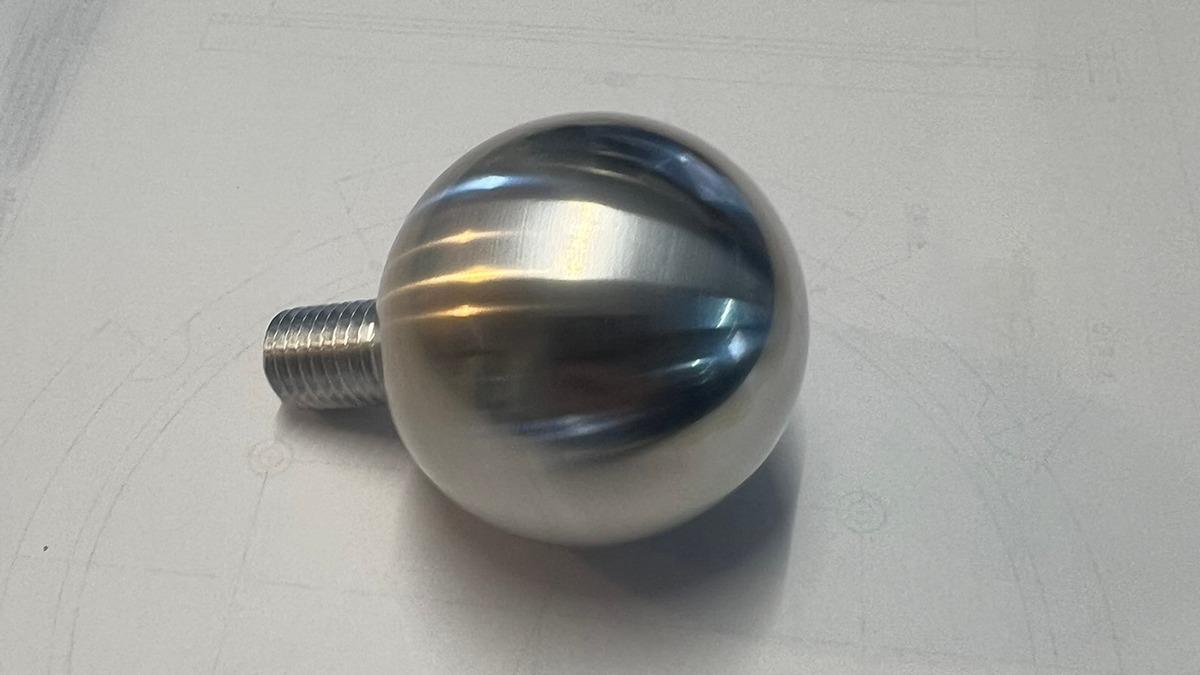 En blank metal kugle med gevind på den ene side, placeret på en hvid overflade.