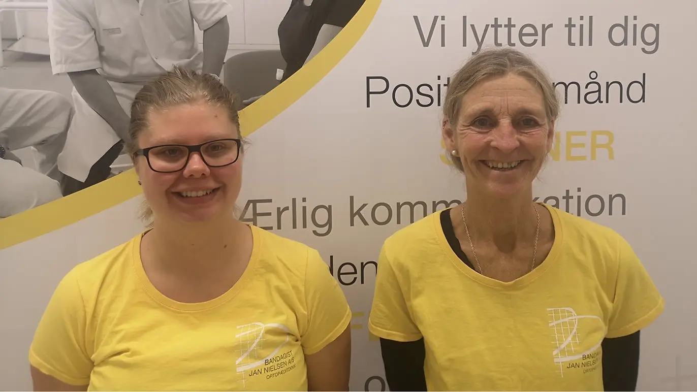 To personer iført gule T-shirts står foran en væg med tekst om kommunikation og lytning.