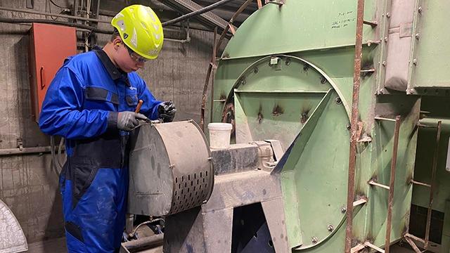 En arbejder i blå overalls og hjelm, der kontrollerer maskineri i en industriel fabrik.