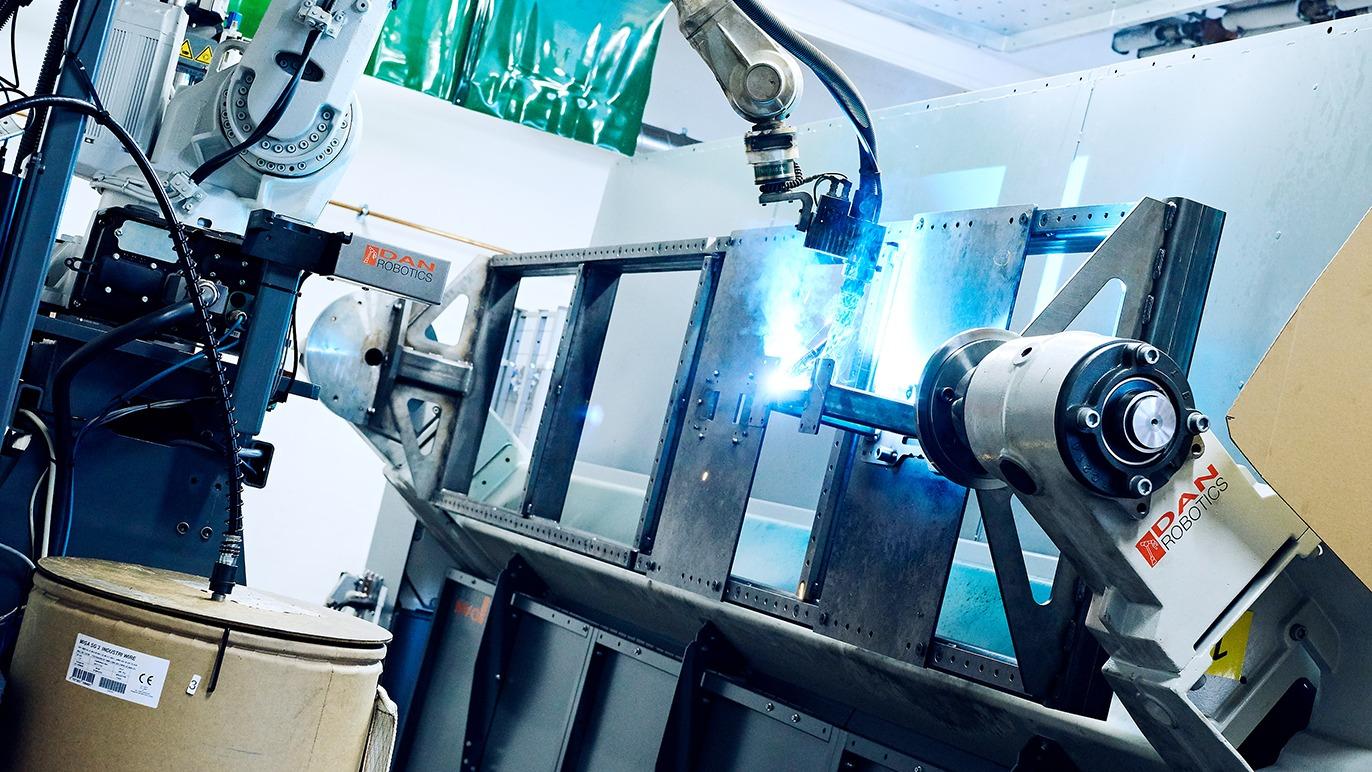 Robotarm svejser metalramme i moderne produktionsmiljø med lys fra svejseprocessen.