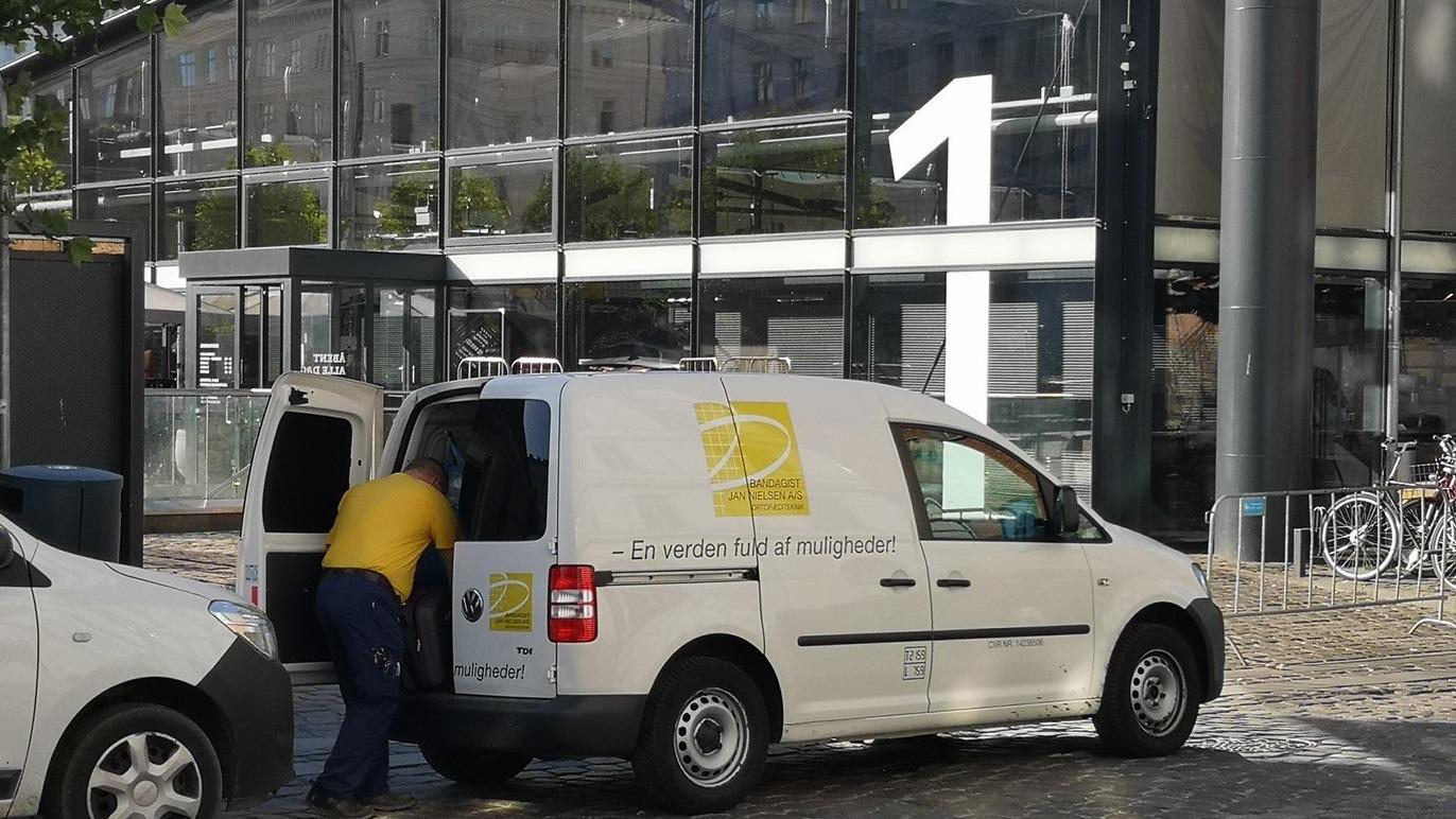 En postbil parkeret foran en bygning med et stort nummer 1, mens en medarbejder læsser varer ind.
