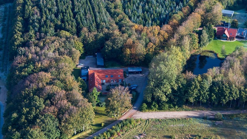 Luftfoto af en gård omgivet af skov, træer og en sø i efterårsfarver.