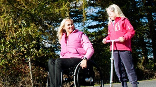 En kvinde i kørestol smiler, mens en lille pige står ved siden af hende med en løbehjul i en solrig park.