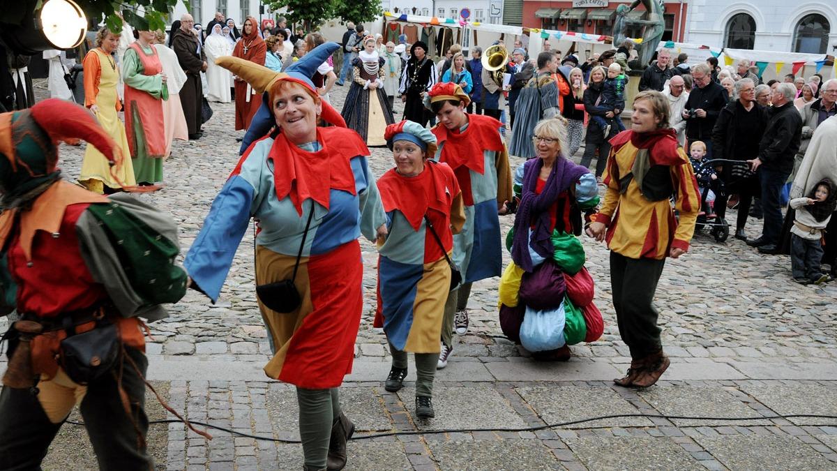 Farverige gøglere i kostumer danser på en brostensbelagt gade, mens publikum ser til.