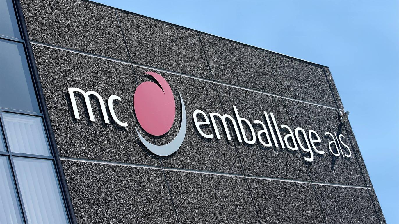 Bygning med skilt for MC Emballage A/S, der viser virksomhedens logo og navn på en grå facade.