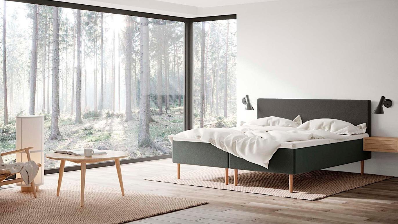 Moderne soveværelse med en dobbeltseng, store vinduer og udsigt til en skov. Møbler i lys træ og minimalistisk design.