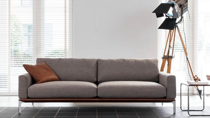 Moderne grå sofa med en brun pude, placeret i et lyst rum med persienner og en lampe på stativ.