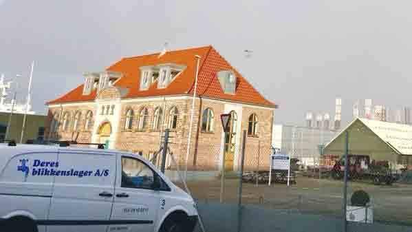 En bygning med rød tag og murstensvægge, parkeret bil foran, i et industriområde med skyer i baggrunden.