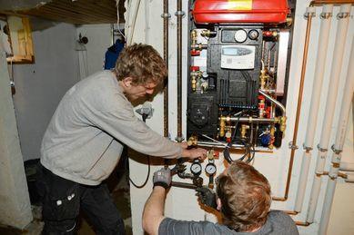 To personer arbejder på en varmepumpe i en kælder, mens de justerer rør og måleinstrumenter.
