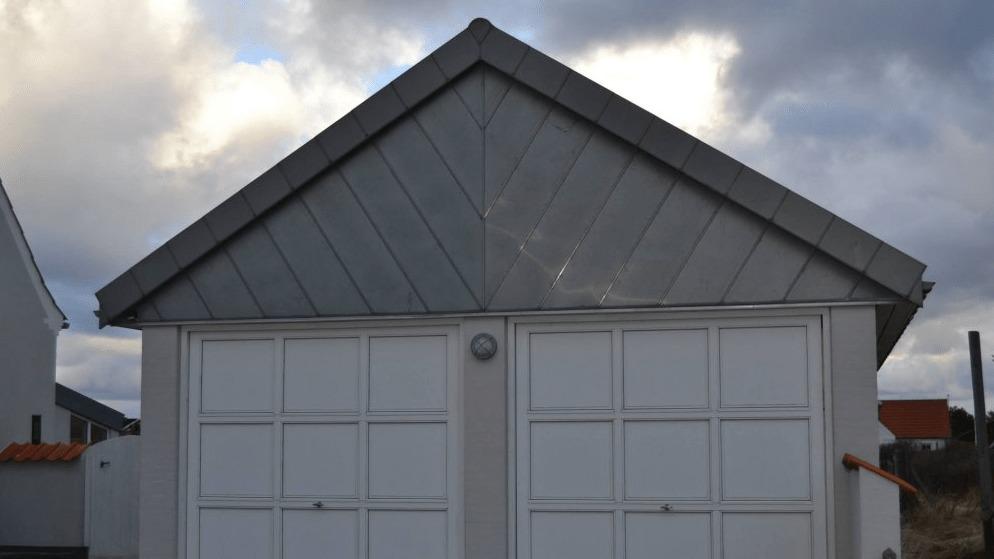 En garage med grå metaltag og hvid garageport, beliggende under en skyet himmel.