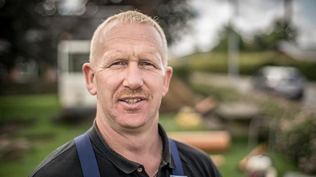 En smilende mand med kort hår og overskæg står foran en havearbejdsplads.