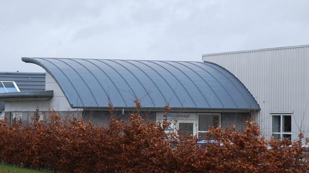Moderne bygning med buet metaltag og brune buske foran.