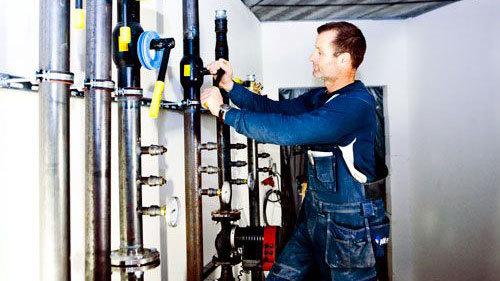 En håndværker arbejder med rørinstallation i et teknikrum med metalrør og værktøj.