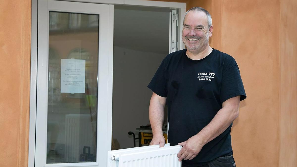 En smilende mand bærer en radiator ved en åben dør i et hus.