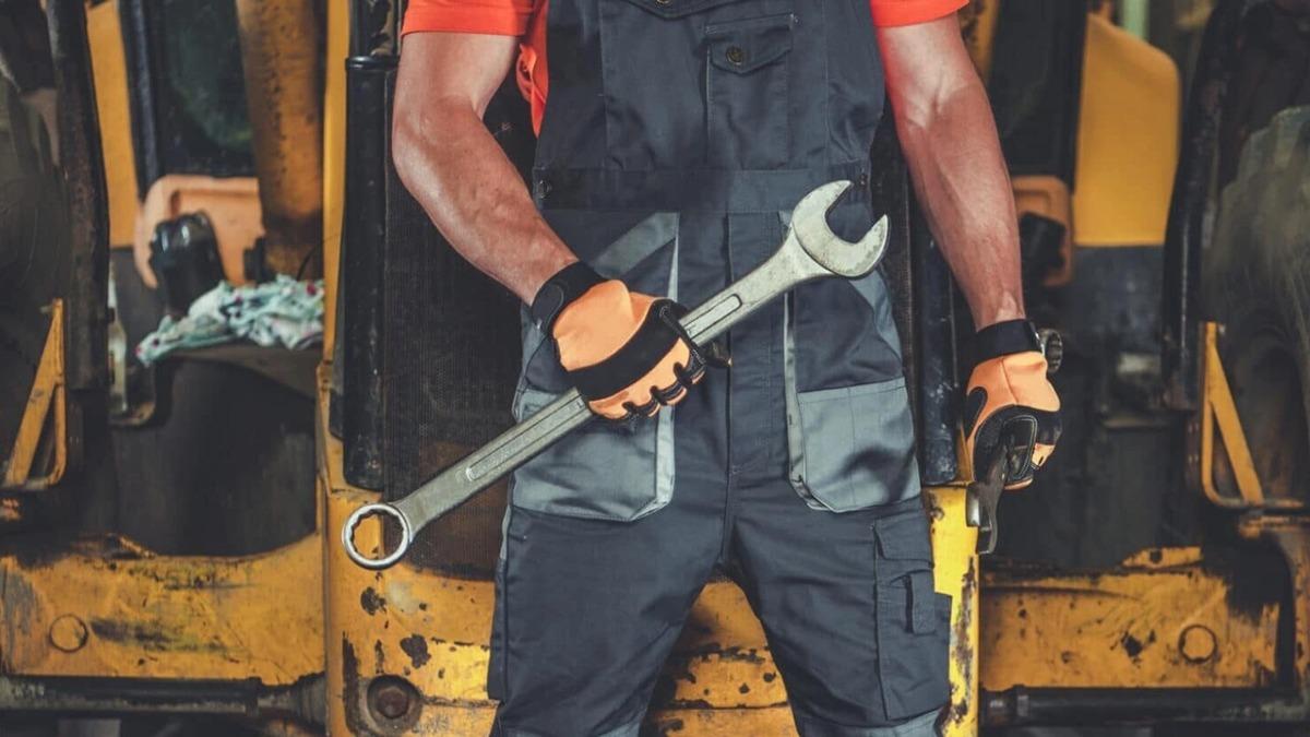 En mekaniker står med en skruenøgle i hånden, iført arbejdstøj og handsker, foran en maskine.