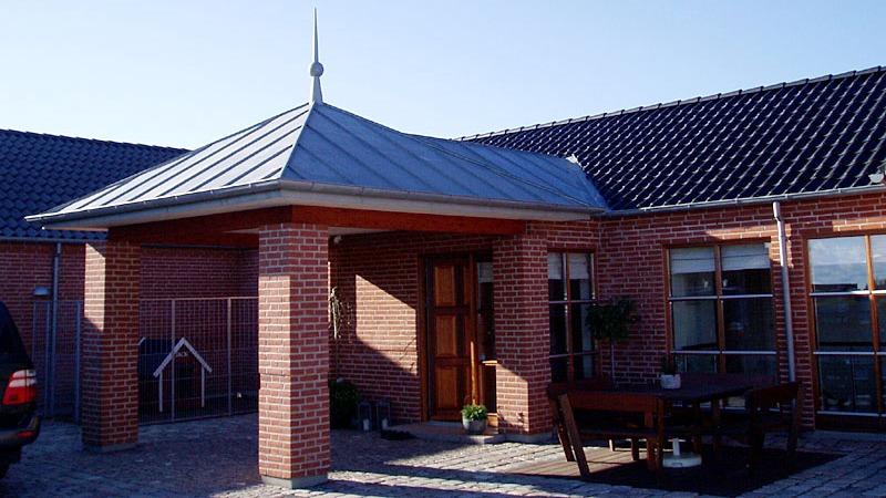 En moderne murstensvilla med en metaltag og en overdækket indgang, omgivet af en stenbelagt gårdsplads.