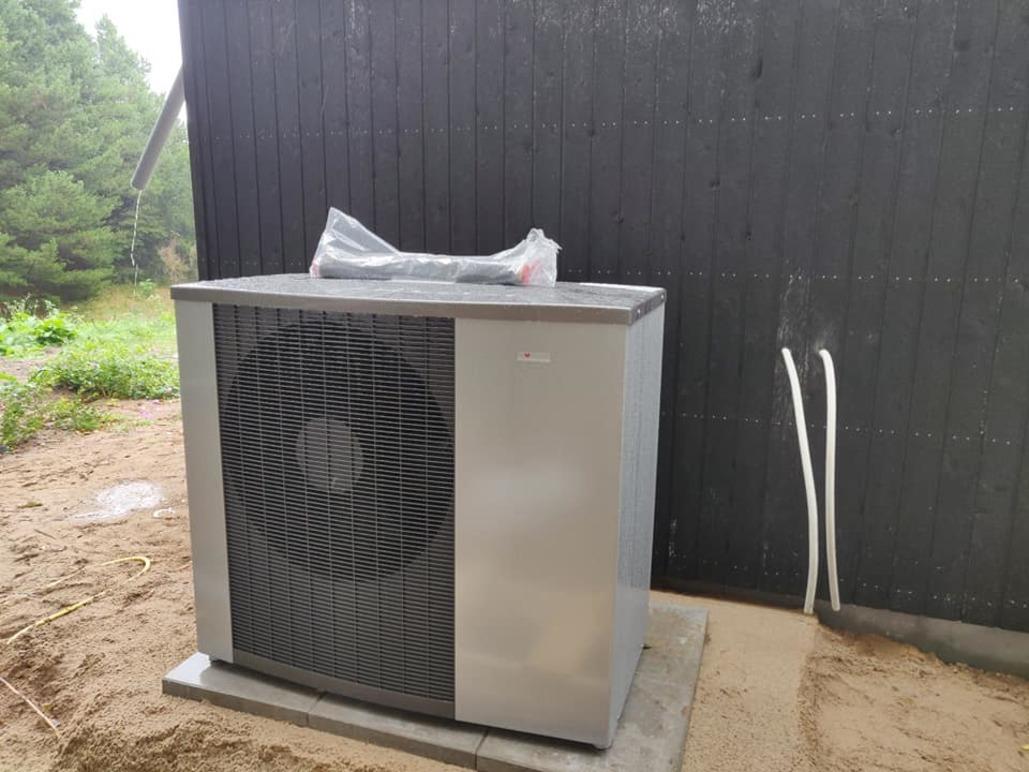 En luftvarmepumpe placeret ved en sort væg, omgivet af sand og græs.