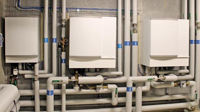 Tre hvide kedler med rørinstallationer i en kælder, der viser et komplekst varmesystem.