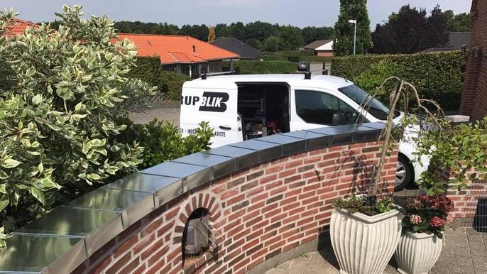 En hvid varevogn med teksten "UPBLIK" parkeret ved en mur med planter og blomster.