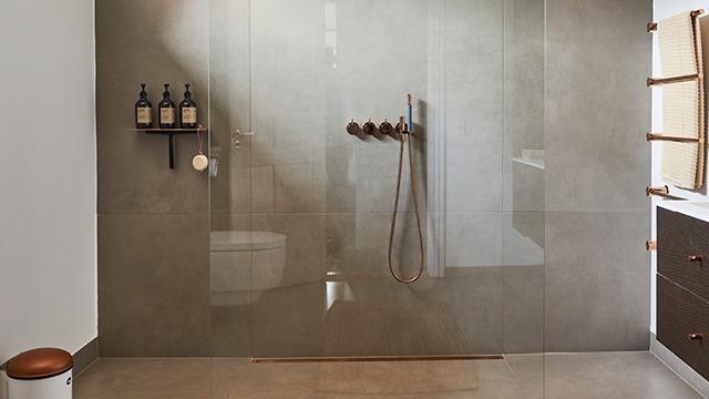 Moderne brusebad med glasvæg, bruserarmatur og hylde til shampooflasker. Minimalistisk indretning i grå nuancer.