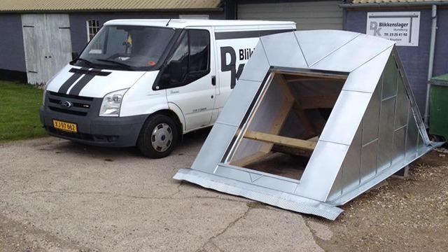 En hvid varevogn parkeret ved siden af en metalbygning med en skrå tagkonstruktion.