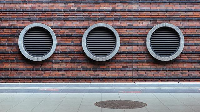 Tre runde ventilationsåbninger i en mur af røde mursten med grå rammer.