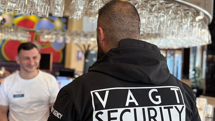 Sikkerhedsvagt i sort jakke med "VAGT SECURITY" står ved baren, mens en bartender smiler.