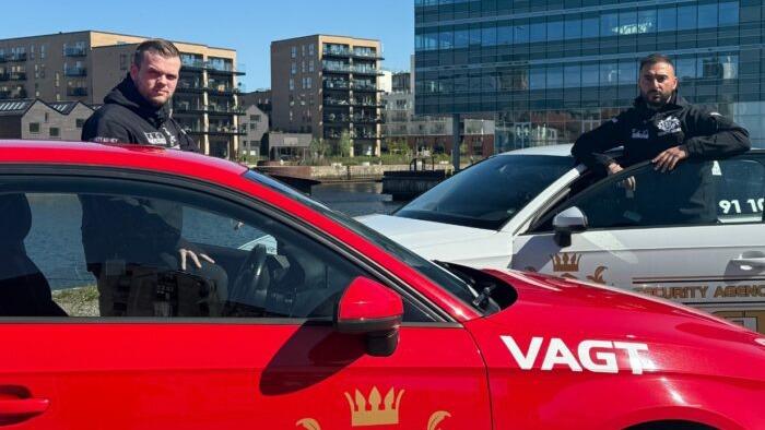 To personer står ved siden af en rød og en hvid bil med sikkerhedsmærker i en bymiljø.