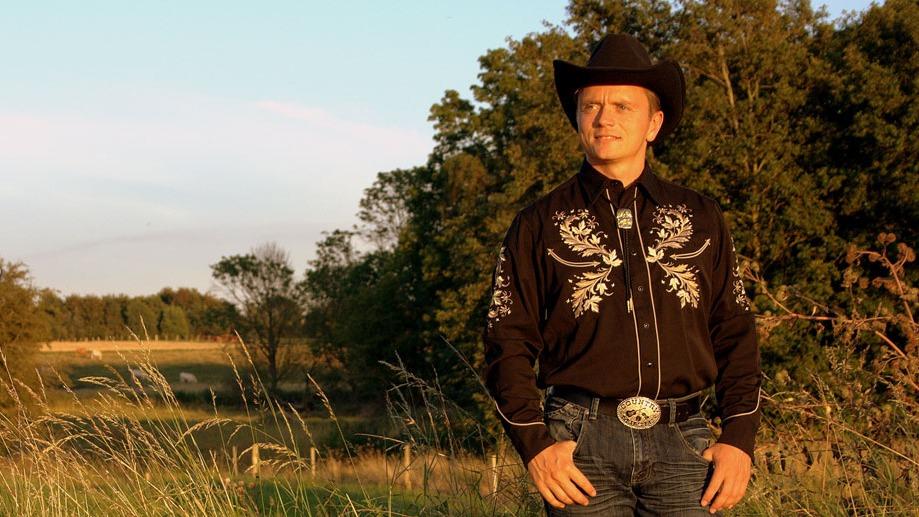 En mand i cowboyhat og broderet skjorte står i en mark med træer i baggrunden under en klar himmel.