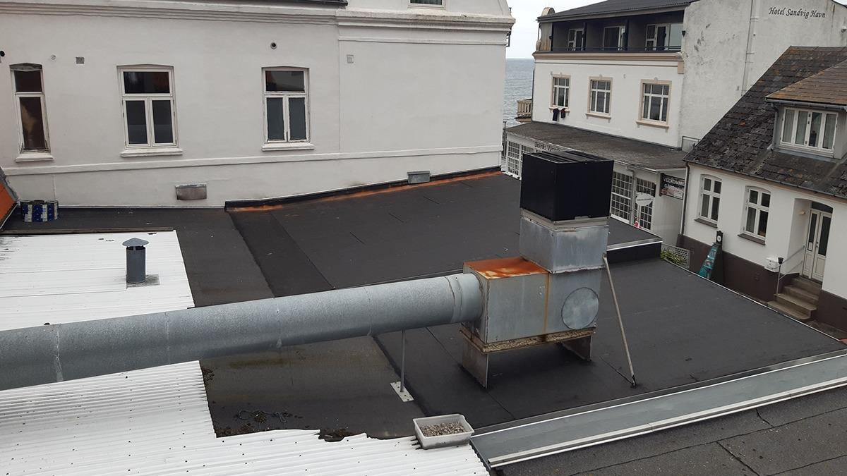 Tag med skorsten og ventilationsrør på en bygning, med udsigt til nabobygninger i baggrunden.