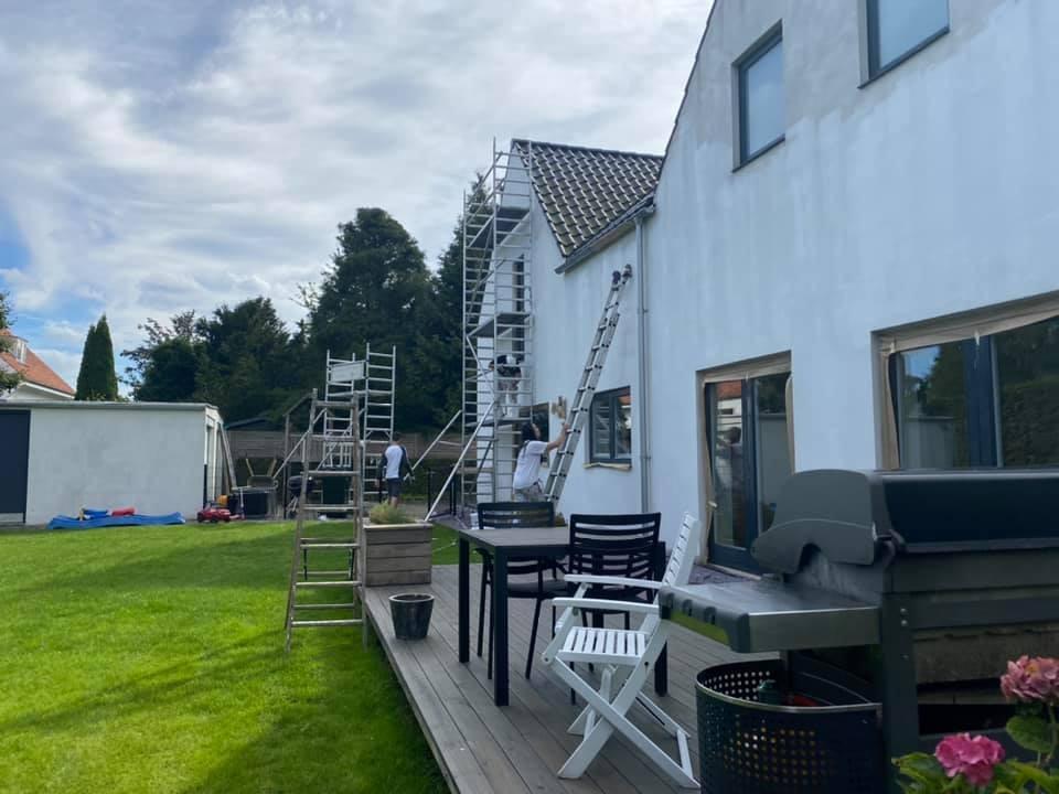 Hus med hvid facade, stillads og have, hvor der er en terrasse med havemøbler og grill.