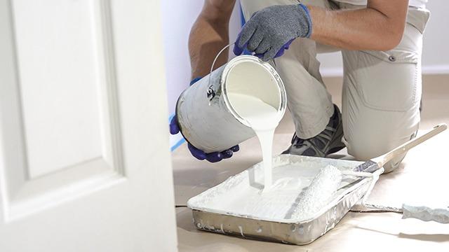 En person hælder hvid maling fra en spand til en malerbakke, mens han står på knæ i et rum.
