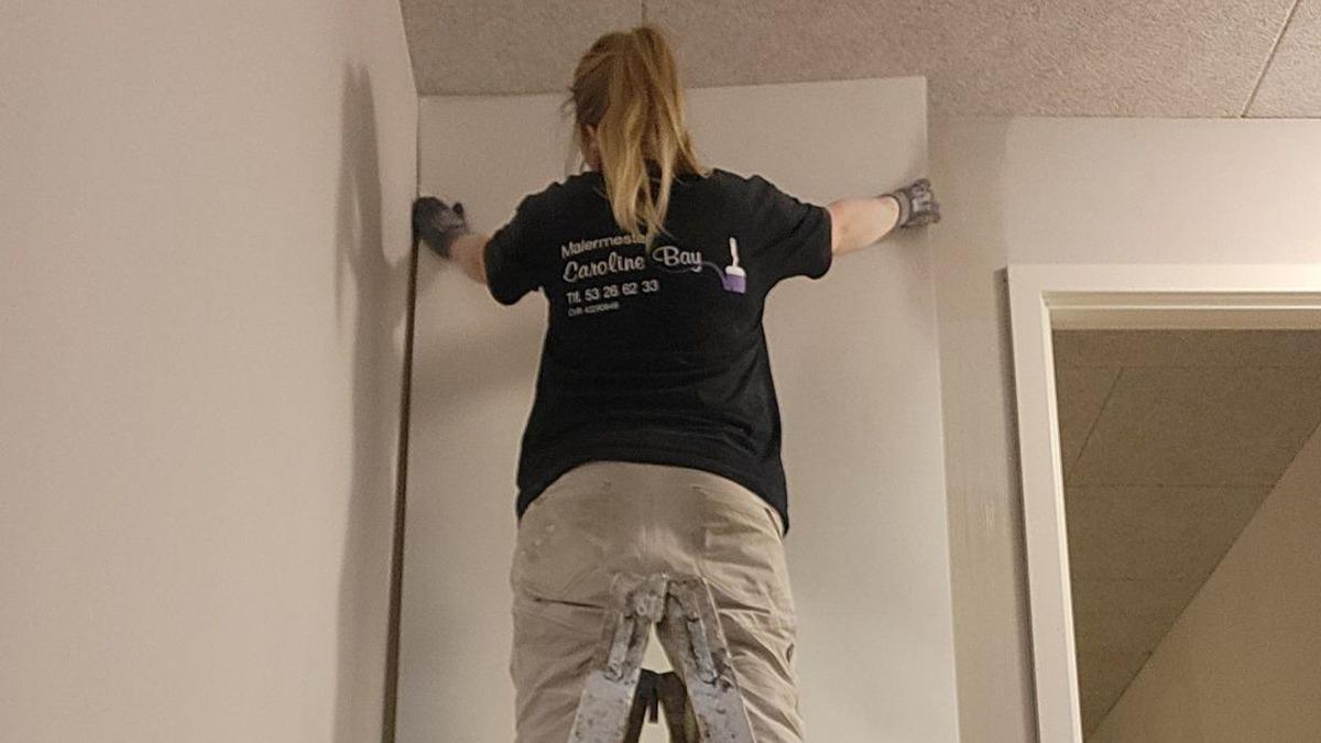 En person maler en væg i et rum, iført en sort t-shirt og arbejdstøj, mens de står på en stige.