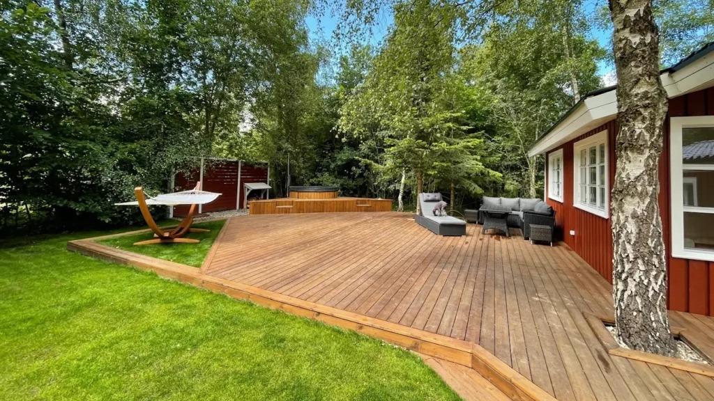 En solrig have med en træterrasse, loungemøbler og en swimmingpool omgivet af grønne træer.