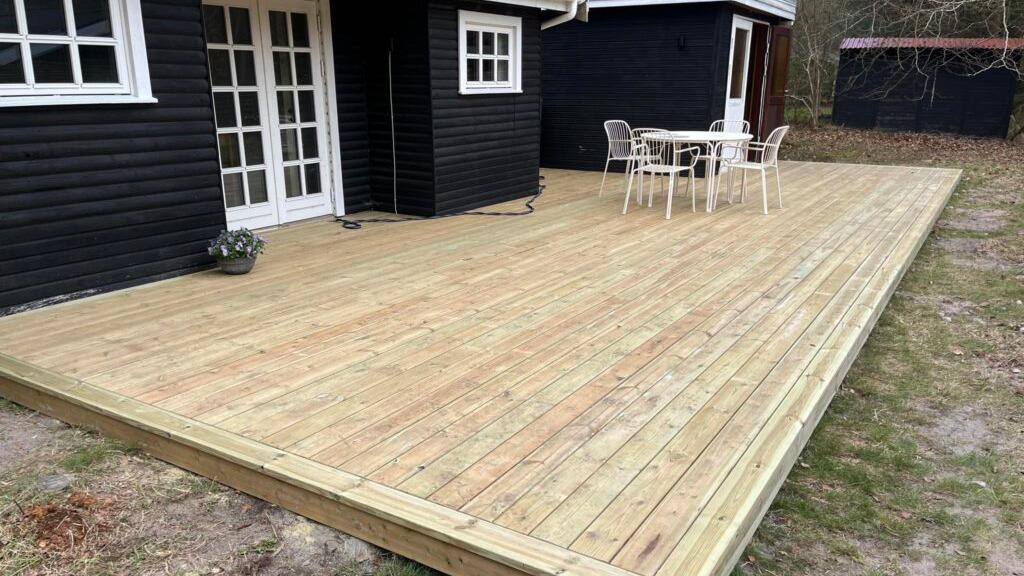 En ny træterrasse foran et sort hus med en hvid dør og en lille spiseplads med borde og stole.