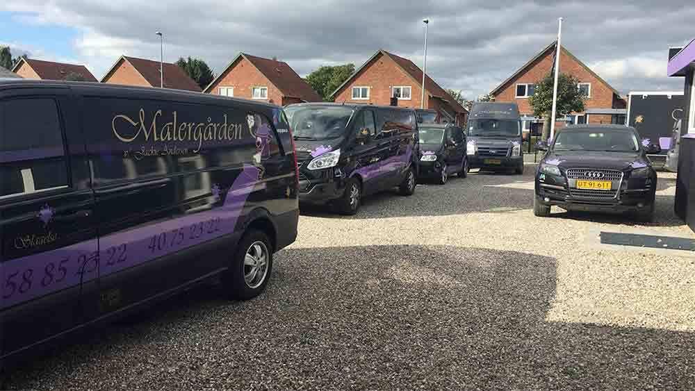 Flere biler og varevogne parkeret på en grusplads foran bygninger i et boligområde.