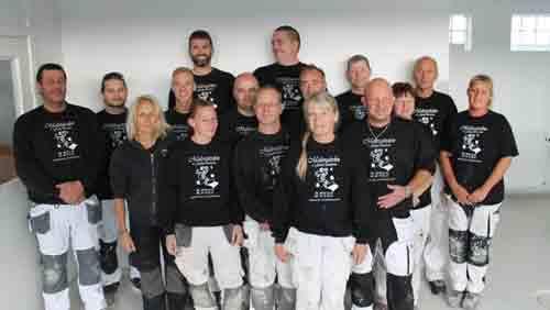 Gruppebillede af 15 personer iført sorte t-shirts og arbejdsbukser, smilende i et lyst rum.