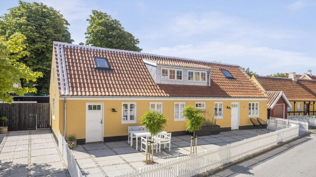 Gul hus med rødt tag og terrasse, omgivet af træer og en hvid stakit i en rolig gade.