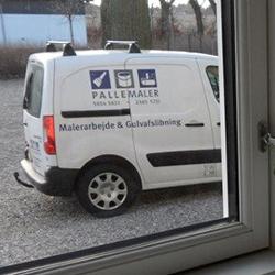 En hvid varevogn med teksten "Pallemaler" parkeret på en grusvej udenfor et vindue.