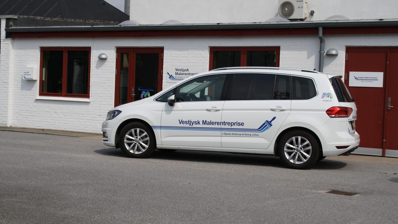 En hvid bil med logo parkeret foran en bygning med røde vinduer og en skilt for "Uddannelses Midtvest".