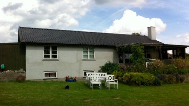 Et hus med græsplæne, hvide havemøbler og en overdækket terrasse under en skyet himmel.