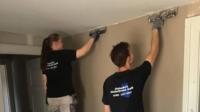 To personer maler en væg indendørs, iført sorte T-shirts med logo og handsker.