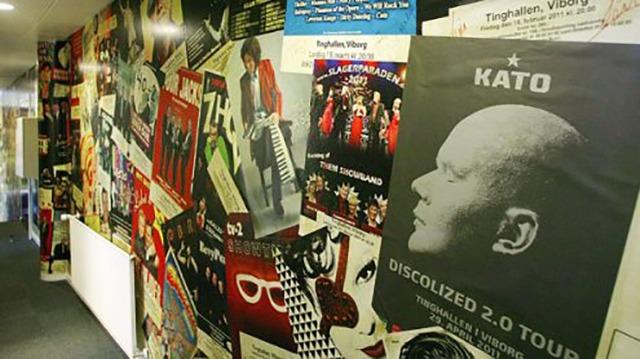 Væggen i en gang med farverige plakater fra koncerter og events, herunder Kato's Discolized 2.0 Tour.