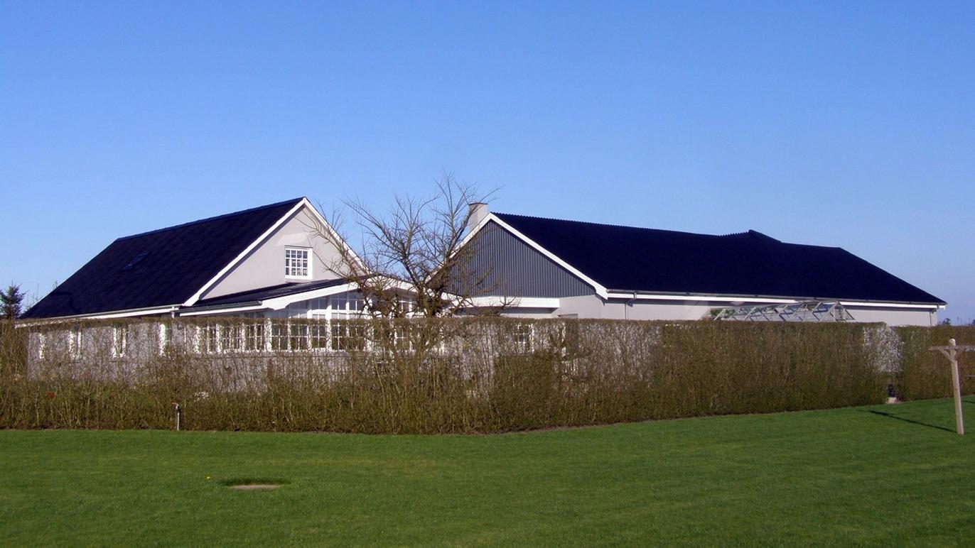 Et moderne hus med sort tag og græsplæne, omgivet af en lav hæk under en klar blå himmel.