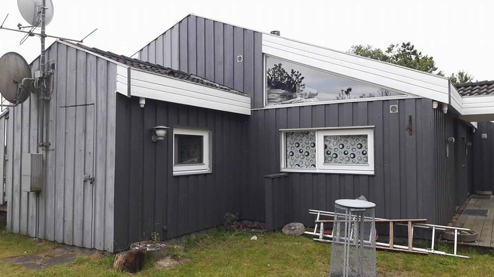 Moderne grå træhus med skrå tag og store vinduer, beliggende i en have med græsplæne og antenner.