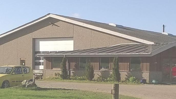 En stor bygning med grå vægge og et fladt tag, omgivet af græs og en gul bil parkeret foran.