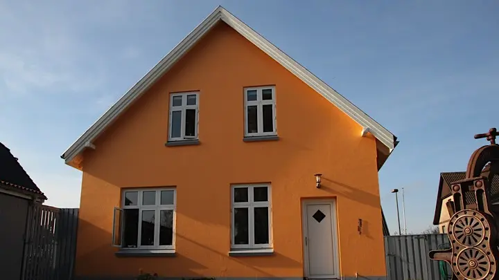 Et gult hus med tre vinduer og en hvid dør under en blå himmel.