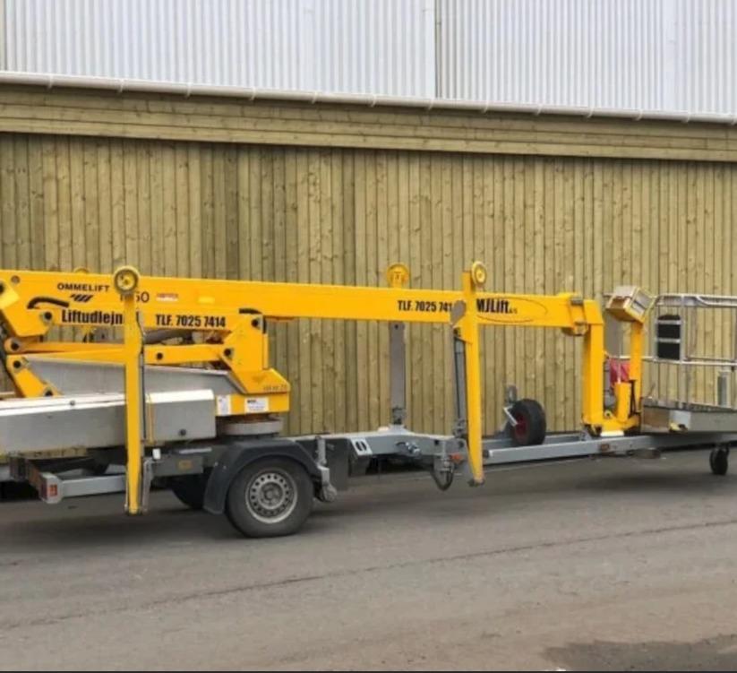 En gul lift med trailer står parkeret ved en trævæg, klar til brug på byggepladsen.