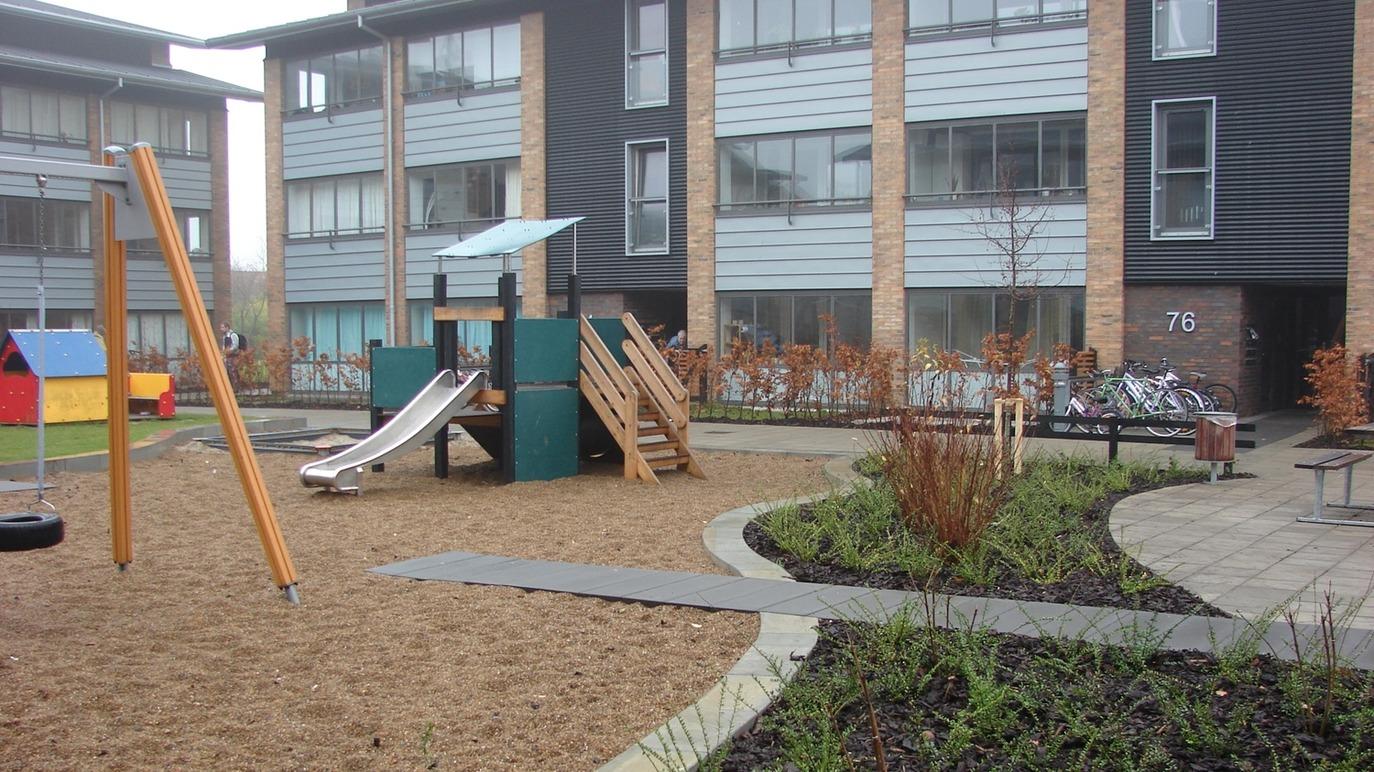 Legestativ med rutsjebane og gynger i en gård mellem moderne bygninger, omgivet af sand og planter.